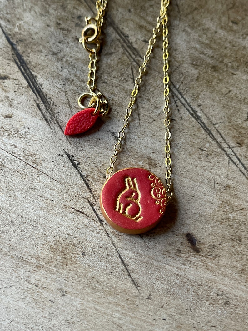 May include: A gold chain necklace with a red circular pendant featuring a gold rabbit silhouette and a gold floral design.