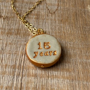 May include: A gold chain necklace with a white ceramic pendant. The pendant has the words "15 years" written in gold.