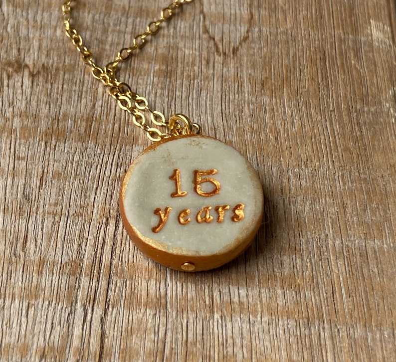 May include: A gold chain necklace with a white ceramic pendant. The pendant has the words "15 years" written in gold.