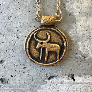 May include: A gold-colored pendant necklace with a circular charm. The charm features a stylized bull or ox figure in a dark bronze color. The necklace chain is a delicate gold color, and the pendant has a textured, antique finish.