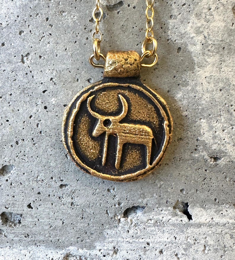 May include: A gold-colored pendant necklace with a circular charm. The charm features a stylized bull or ox figure in a dark bronze color. The necklace chain is a delicate gold color, and the pendant has a textured, antique finish.