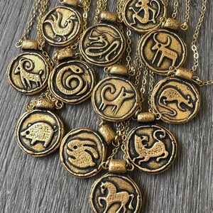 May include: Gold-colored pendant necklaces with various zodiac animal symbols. Each circular pendant features a raised black outline of an animal, such as a horse, monkey, or pig, on a gold background. The necklaces are displayed on a wooden surface.