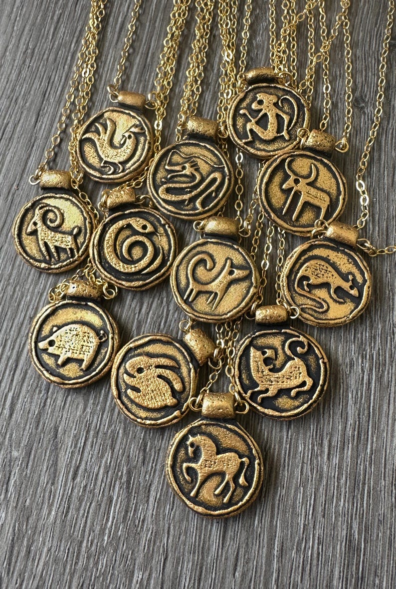 May include: Gold-colored pendant necklaces with various zodiac animal symbols. Each circular pendant features a raised black outline of an animal, such as a horse, monkey, or pig, on a gold background. The necklaces are displayed on a wooden surface.