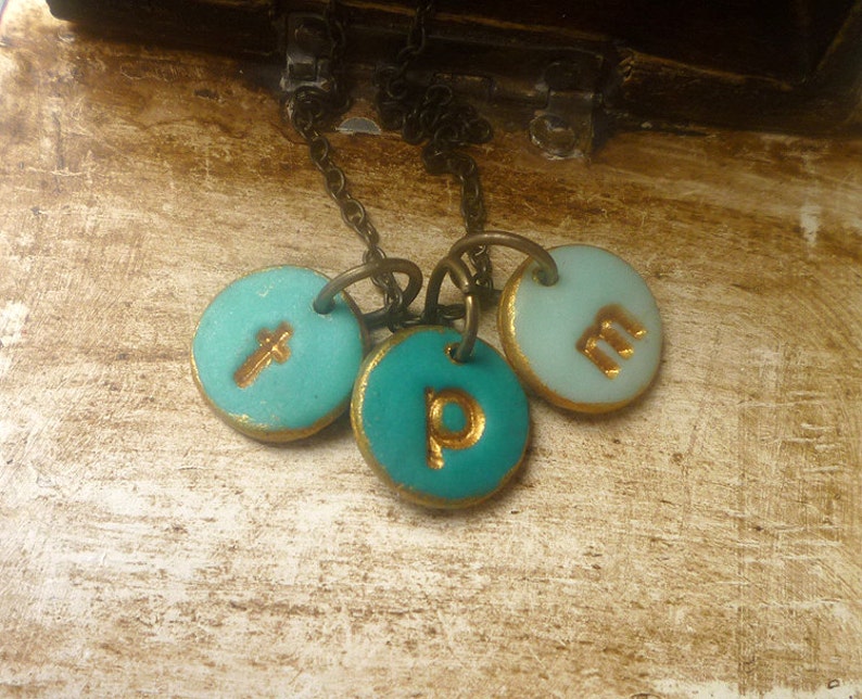 May include: A turquoise and gold necklace with three round charms. Each charm has a gold letter on it: T, P, and M.