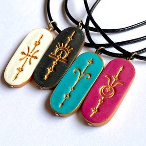 May include: Four pendant necklaces with black cords. Each pendant is a different color: white, black, teal, and magenta. Each pendant has a gold-colored design. The pendants are oval-shaped with gold-colored edges.