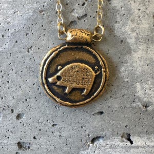 May include: A gold-colored pendant necklace featuring a circular charm with a detailed boar design. The boar is in the center of the charm, which is framed by a dark border. The necklace chain is also gold.