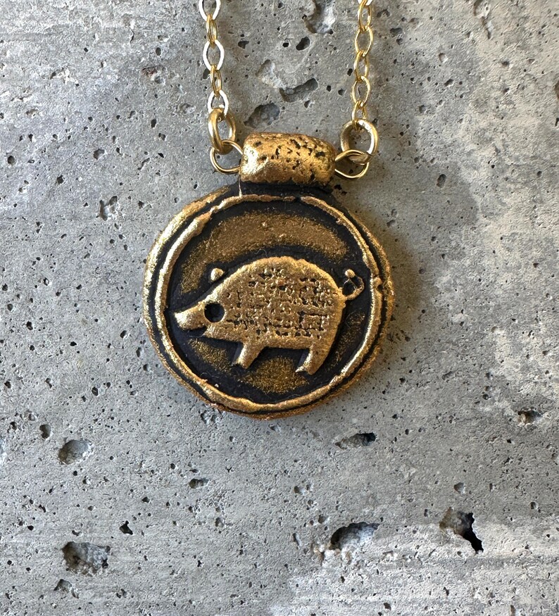 May include: A gold-colored pendant necklace featuring a circular charm with a detailed boar design. The boar is in the center of the charm, which is framed by a dark border. The necklace chain is also gold.