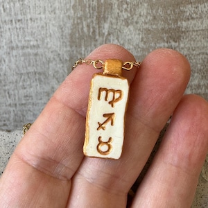 May include: A gold-colored necklace featuring a rectangular pendant with a white background and gold trim. The pendant displays three zodiac symbols in brown: Virgo, Sagittarius, and Taurus. The necklace is held by a hand.