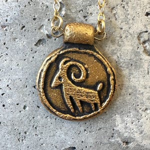 May include: A gold-colored pendant necklace featuring a circular charm with a ram design. The ram is in profile, with a curved horn and detailed body. The pendant hangs from a delicate gold chain, creating a vintage aesthetic.