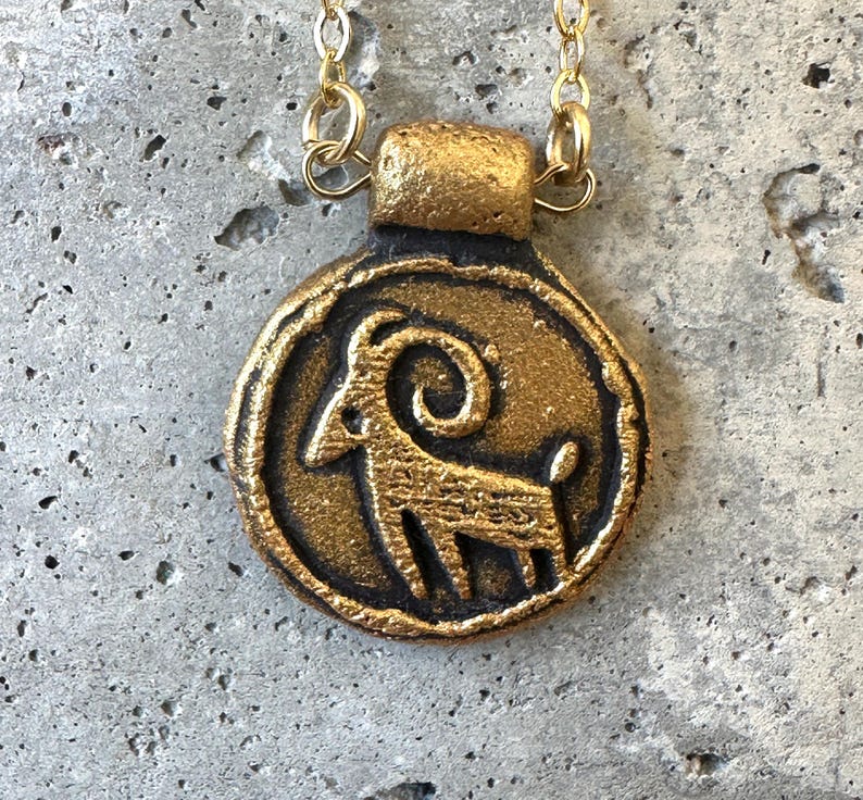 May include: A gold-colored pendant necklace featuring a circular charm with a ram design. The ram is in profile, with a curved horn and detailed body. The pendant hangs from a delicate gold chain, creating a vintage aesthetic.