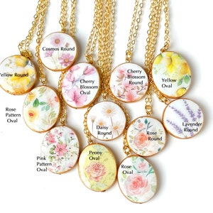 May include: A collection of 10 floral-themed necklaces with gold chains. The pendants are round and oval, featuring various floral designs, including roses, daisies, cherry blossoms, and lavender. Each pendant has a different floral name and shape written on it.