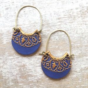 May include: A pair of gold hoop earrings with a blue ceramic crescent moon pendant. The pendant features a gold filigree design.