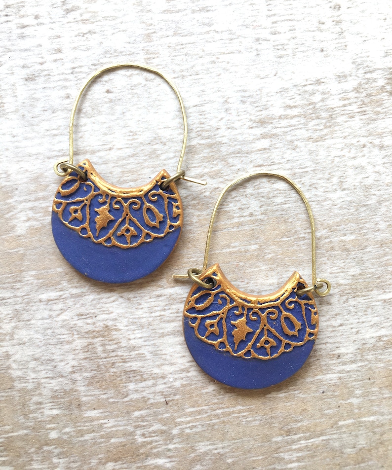 May include: A pair of gold hoop earrings with a blue ceramic crescent moon pendant. The pendant features a gold filigree design.