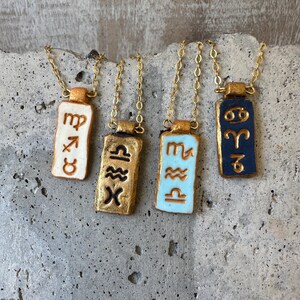 May include: Four gold-toned necklaces with rectangular pendants featuring zodiac symbols. The pendants are painted in various colors, including white, gold, light blue, and navy blue. Each pendant hangs from a delicate gold chain.