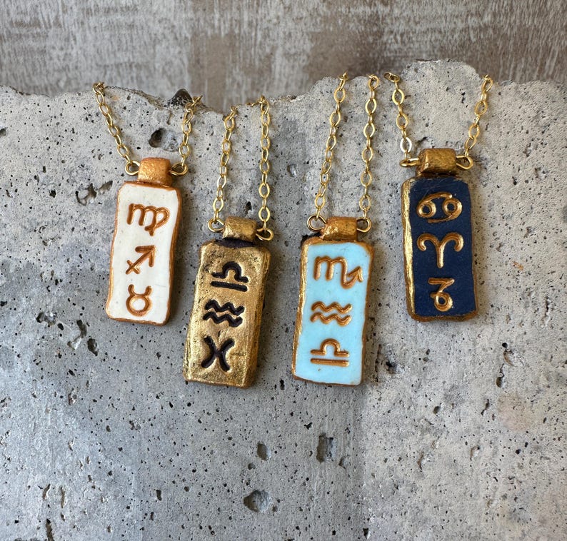May include: Four gold-toned necklaces with rectangular pendants featuring zodiac symbols. The pendants are painted in various colors, including white, gold, light blue, and navy blue. Each pendant hangs from a delicate gold chain.