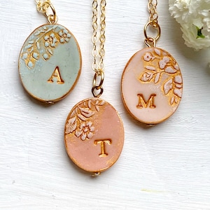 May include: Three gold chain necklaces with oval pendants. Each pendant has a floral design and a different letter: A, M, and T. The pendants are light blue, pink, and peach.