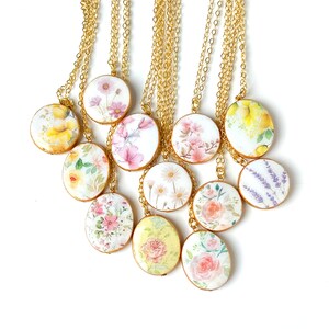 May include: A set of 10 gold chain necklaces with oval-shaped pendants featuring different floral patterns. The pendants are white with watercolour-style floral designs in shades of pink, yellow, and purple.