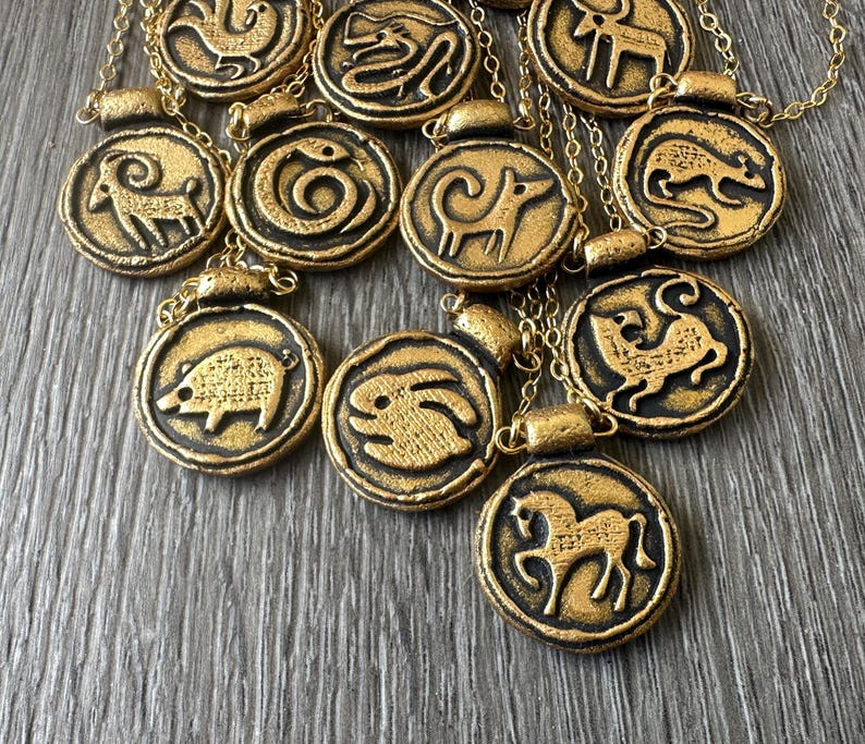 May include: Gold-colored pendant necklaces featuring zodiac animal symbols. Each circular pendant has a black border and a gold-colored animal design. The necklaces are on gold chains.