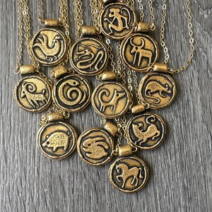 May include: Gold-toned pendant necklaces featuring zodiac animal symbols. Each circular pendant has a raised relief design of a different animal, with a dark border. The necklaces are on gold chains, and are displayed on a gray wooden surface.