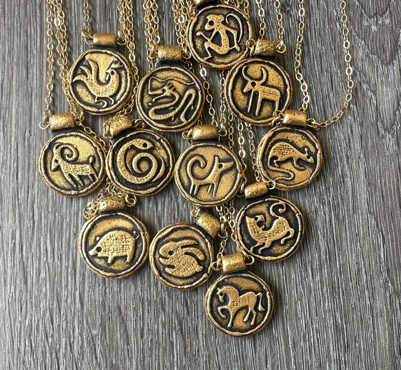 May include: Gold-toned pendant necklaces featuring zodiac animal symbols. Each circular pendant has a raised relief design of a different animal, with a dark border. The necklaces are on gold chains, and are displayed on a gray wooden surface.