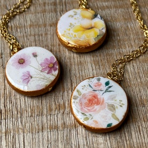 May include: Three gold necklaces with round pendants featuring floral designs. The pendants have a white background with watercolour-style flowers in shades of pink, yellow, and orange.