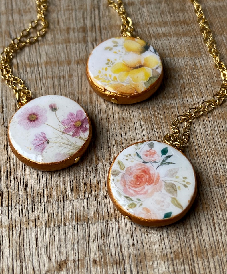 May include: Three gold necklaces with round pendants featuring floral designs. The pendants have a white background with watercolour-style flowers in shades of pink, yellow, and orange.