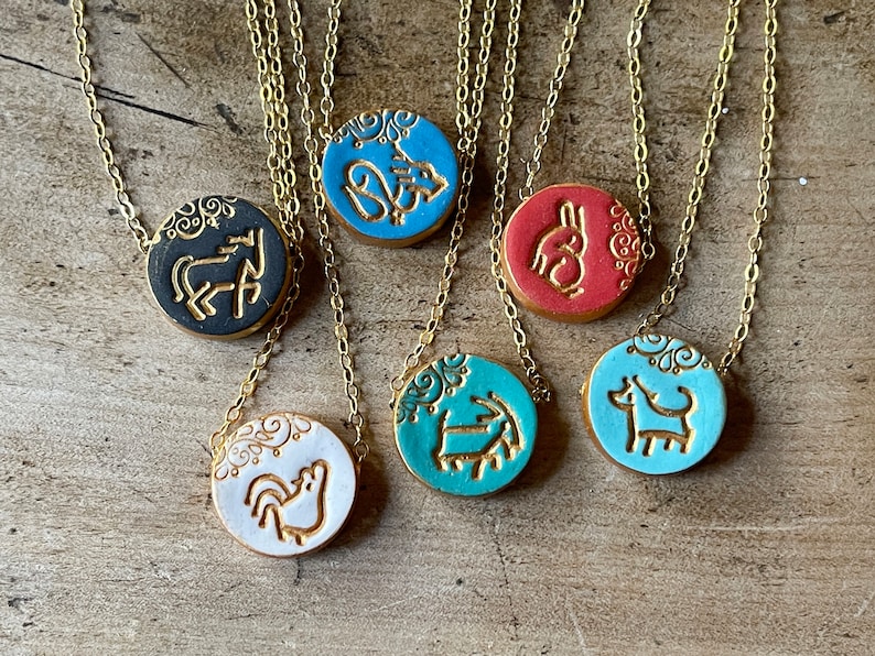 May include: Six gold chain necklaces with round ceramic pendants featuring different animal designs. The pendants are in various colors, including black, white, blue, and red.
