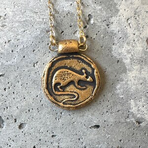 May include: A gold-colored pendant necklace featuring a circular charm with a detailed rat design. The rat is depicted in a side profile, set against a dark background. The necklace has a delicate gold chain.