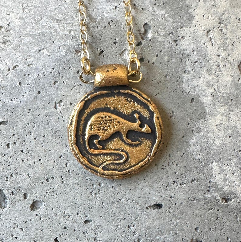 May include: A gold-colored pendant necklace featuring a circular charm with a detailed rat design. The rat is depicted in a side profile, set against a dark background. The necklace has a delicate gold chain.