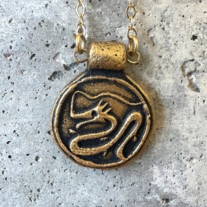 May include: A gold-colored necklace with a circular pendant featuring a black dragon design. The pendant is attached to a delicate gold chain. The dragon is depicted in a stylized, winding form within the circle.