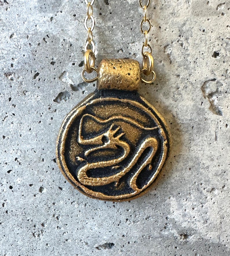 May include: A gold-colored necklace with a circular pendant featuring a black dragon design. The pendant is attached to a delicate gold chain. The dragon is depicted in a stylized, winding form within the circle.