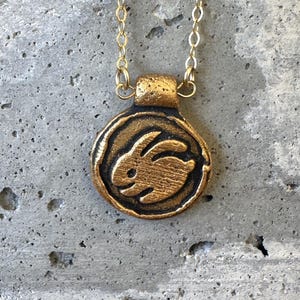 May include: A gold-colored pendant necklace featuring a circular charm with a raised image of a rabbit. The rabbit is in the center of the charm, which has a textured border. The necklace chain is also gold-colored.