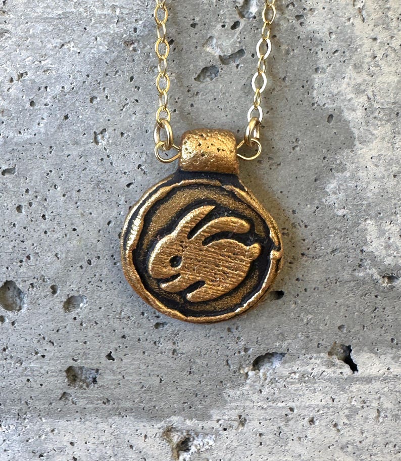 May include: A gold-colored pendant necklace featuring a circular charm with a raised image of a rabbit. The rabbit is in the center of the charm, which has a textured border. The necklace chain is also gold-colored.