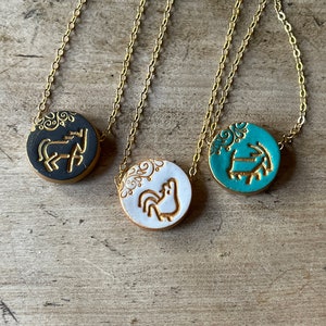 May include: Three gold chain necklaces with round ceramic pendants. The pendants feature a rooster, a horse, and a ram, all in gold on a colored background. The backgrounds are black, white, and teal.