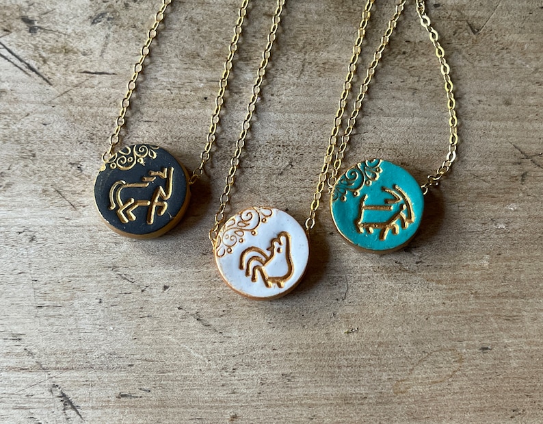 May include: Three gold chain necklaces with round ceramic pendants. The pendants feature a rooster, a horse, and a ram, all in gold on a colored background. The backgrounds are black, white, and teal.