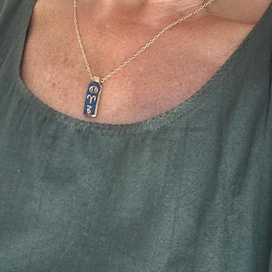 May include: A gold-colored chain necklace with a rectangular pendant. The pendant is navy blue with gold-colored symbols. The necklace is worn against a green top.