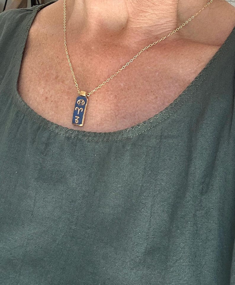 May include: A gold-colored chain necklace with a rectangular pendant. The pendant is navy blue with gold-colored symbols. The necklace is worn against a green top.