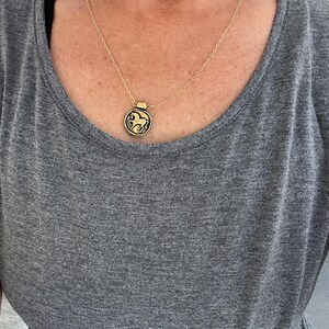 May include: Gold-toned necklace with a circular pendant featuring a detailed horse design. The pendant hangs from a delicate chain. The necklace is worn over a gray t-shirt.