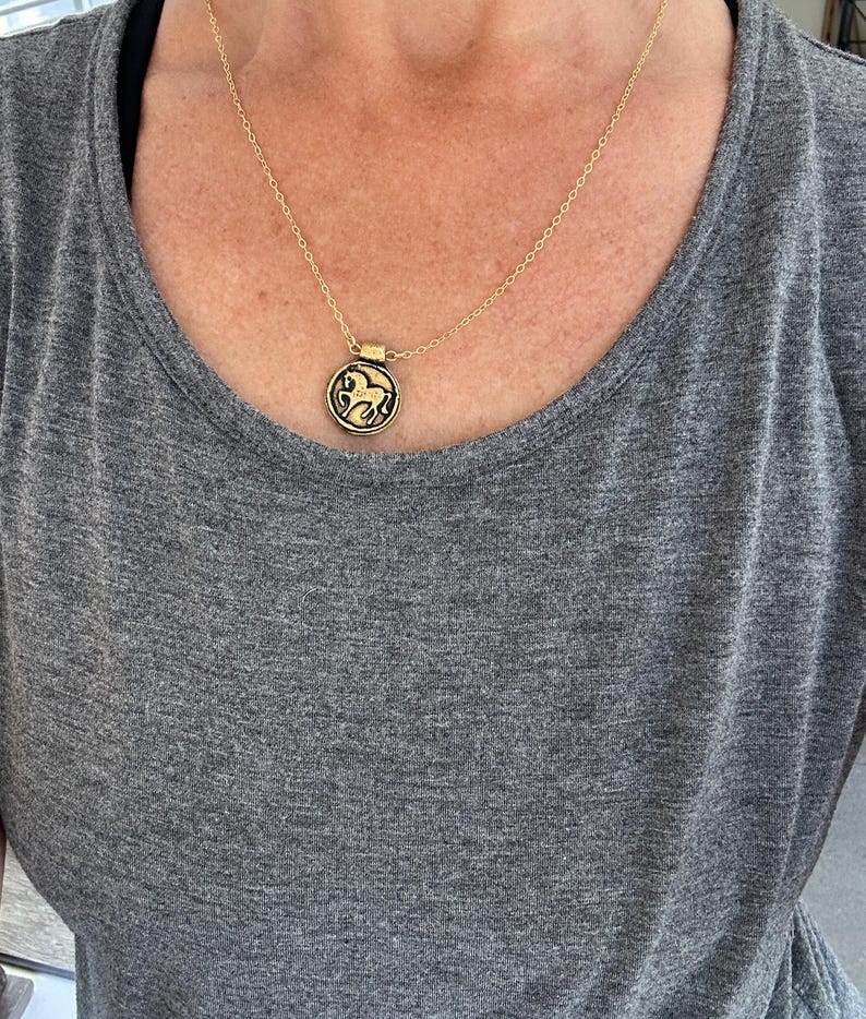May include: Gold-toned necklace with a circular pendant featuring a detailed horse design. The pendant hangs from a delicate chain. The necklace is worn over a gray t-shirt.