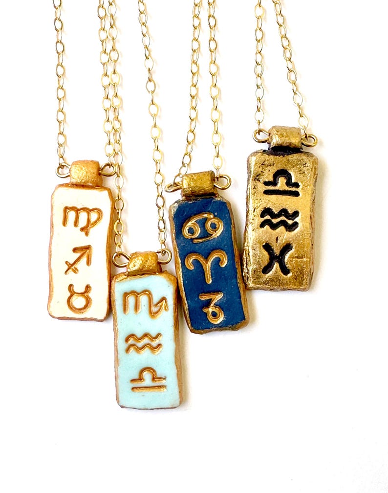 May include: Four gold-colored chain necklaces with rectangular pendants. Each pendant features a different zodiac symbol in gold against a background of white, light blue, or dark blue. The pendants have a textured, metallic appearance.