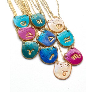 K&ouml;nnte beinhalten: Ein Set aus 12 goldenen Ketten mit Keramik-Anh&auml;ngern, die die astrologischen Zeichen zeigen. Jeder Anh&auml;nger hat eine andere Farbe, darunter T&uuml;rkis, Blau, Gr&uuml;n, Pink und Wei&szlig;.