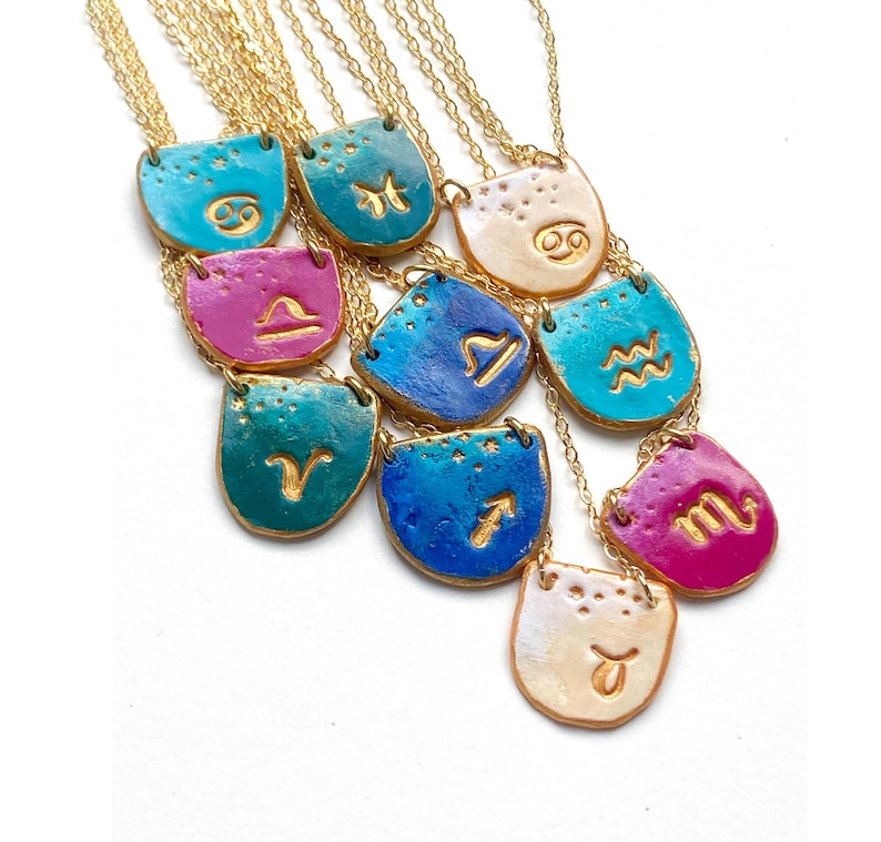 K&ouml;nnte beinhalten: Ein Set aus 12 goldenen Ketten mit Keramik-Anh&auml;ngern, die die astrologischen Zeichen zeigen. Jeder Anh&auml;nger hat eine andere Farbe, darunter T&uuml;rkis, Blau, Gr&uuml;n, Pink und Wei&szlig;.