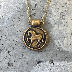 May include: A gold-colored pendant necklace featuring a circular charm with a horse silhouette. The horse is depicted in a side profile within a raised border. The chain and pendant are made of a gold-toned metal, creating a vintage aesthetic.