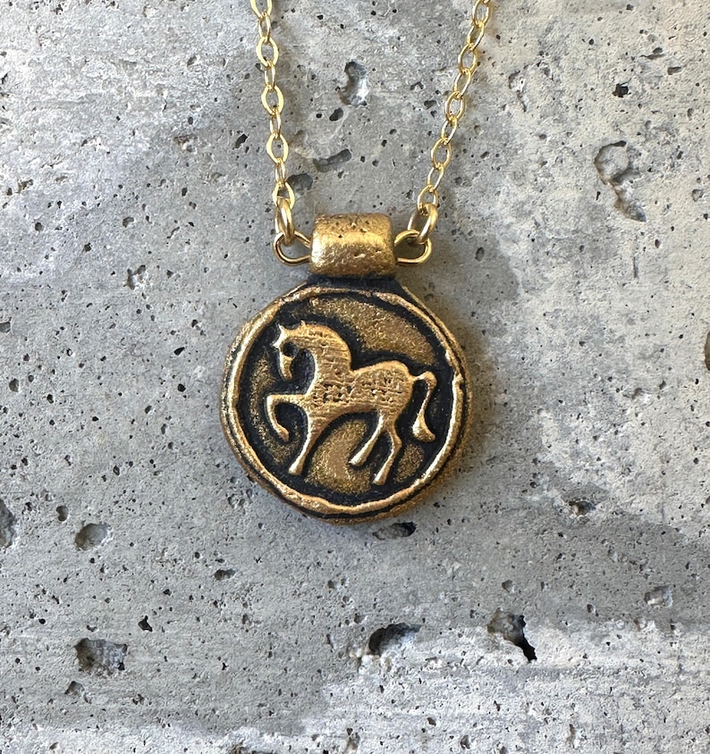 May include: A gold-colored pendant necklace featuring a circular charm with a horse silhouette. The horse is depicted in a side profile within a raised border. The chain and pendant are made of a gold-toned metal, creating a vintage aesthetic.