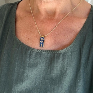 May include: A gold-colored chain necklace with a rectangular, dark blue pendant. The pendant features gold-colored zodiac symbols. The necklace is worn against a green top.
