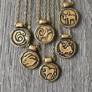 May include: Gold-colored pendant necklaces with various animal designs, including a snake, ram, rabbit, horse, and bull. The pendants are circular with a raised design and are suspended from delicate gold chains. The necklaces are displayed on a gray wooden surface.