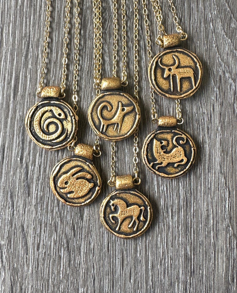 May include: Gold-colored pendant necklaces with various animal designs, including a snake, ram, rabbit, horse, and bull. The pendants are circular with a raised design and are suspended from delicate gold chains. The necklaces are displayed on a gray wooden surface.