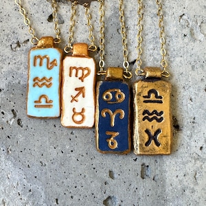 May include: Four gold-colored necklaces with rectangular pendants. Each pendant features a different zodiac symbol in relief, with backgrounds in light blue, white, navy blue, and gold. The chains are delicate and gold-toned.