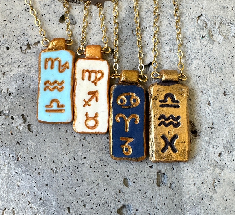 May include: Four gold-colored necklaces with rectangular pendants. Each pendant features a different zodiac symbol in relief, with backgrounds in light blue, white, navy blue, and gold. The chains are delicate and gold-toned.