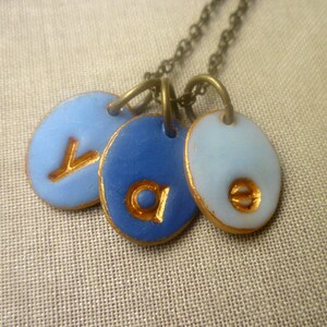May include: A gold chain necklace with three blue and white ceramic pendants. Each pendant has a gold letter on it, spelling out the word "Ava".